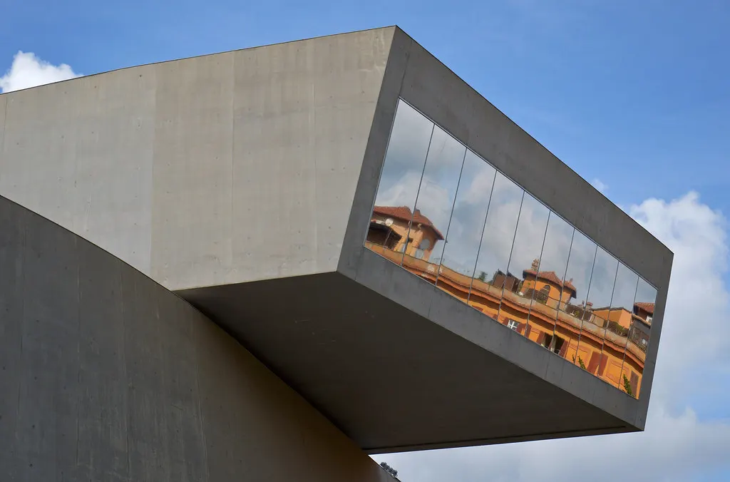 Uitkragende betonnen gevel van het MAXXI museum met glazen raam dat oranje gebouwen weerspiegelt