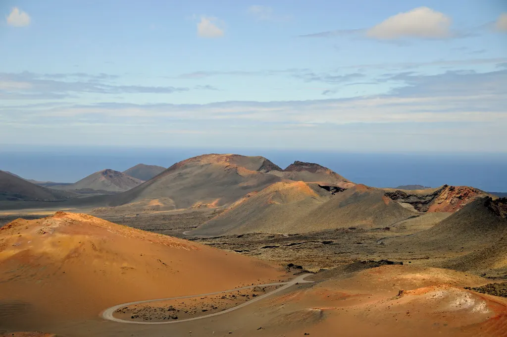 Rood-oranje vulkaankegels en een kronkelende weg in Timanfaya, met de zee en blauwe lucht op de achtergrond