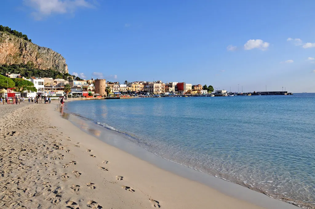 Zandstrand van Mondello met voetsporen, helderblauwe zee, kleurrijke huizen en een rotswand