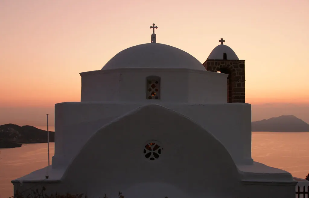Witte kerk met koepel en klokkentoren in Plaka, silhouet tegen een roze zonsonderganghemel boven de zee