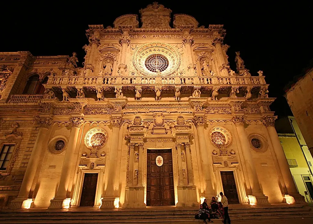 Verlichte barokke gevel van de Basilica di Santa Croce in Lecce bij nacht, met roosvenster en rijk beeldhouwwerk