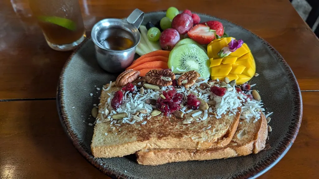 Wentelteefjes met kokos, pecannoten en cranberry's naast vers fruit en een kannetje siroop op een houten tafel