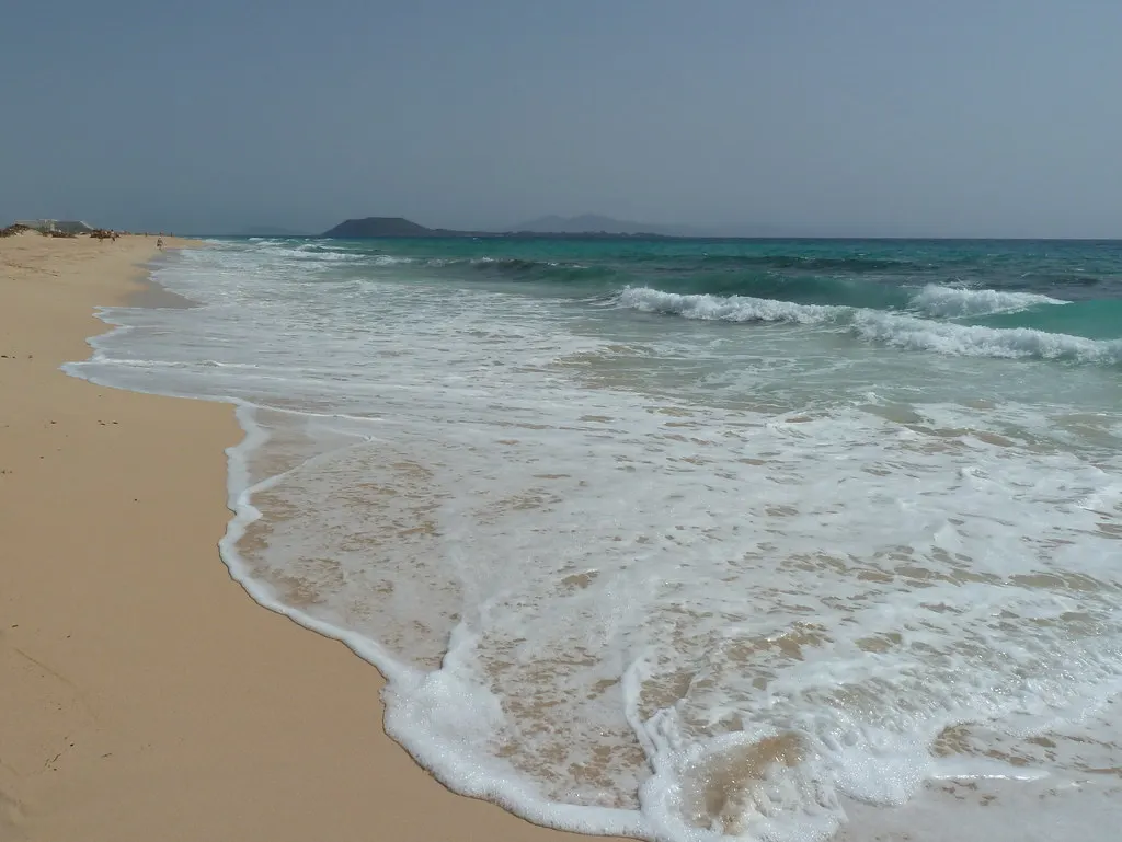 Zandstrand met golven en turquoise zee bij Corralejo, met het eiland Lobos aan de horizon