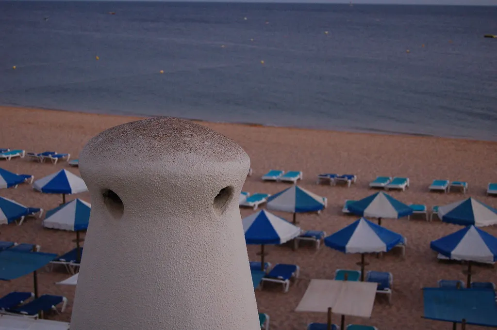 Uitzicht over strand met blauwe parasols en ligbedden naar de zee, met een witte Algarve-schoorsteen op de voorgrond