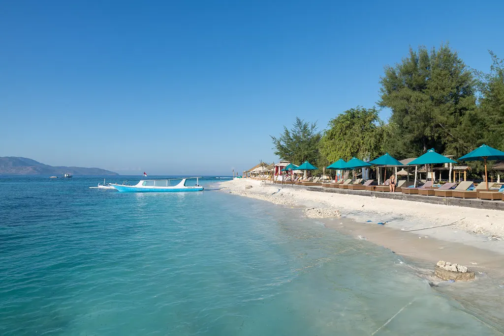 Wit zandstrand van Gili Trawangan met turquoise water, een boot en strandbedjes onder groene parasols
