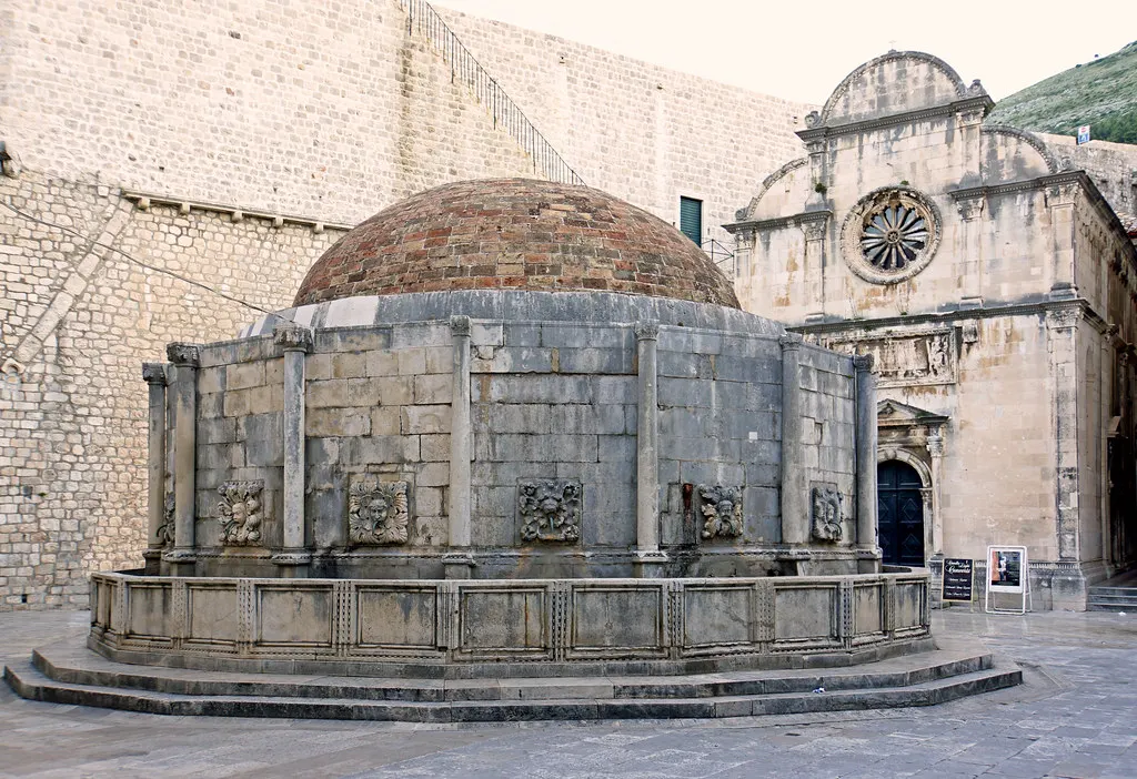 De Grote Onofrio-fontein met koepel en stenen maskers, met daarachter de gevel van de St. Saviour-kerk in Dubrovnik