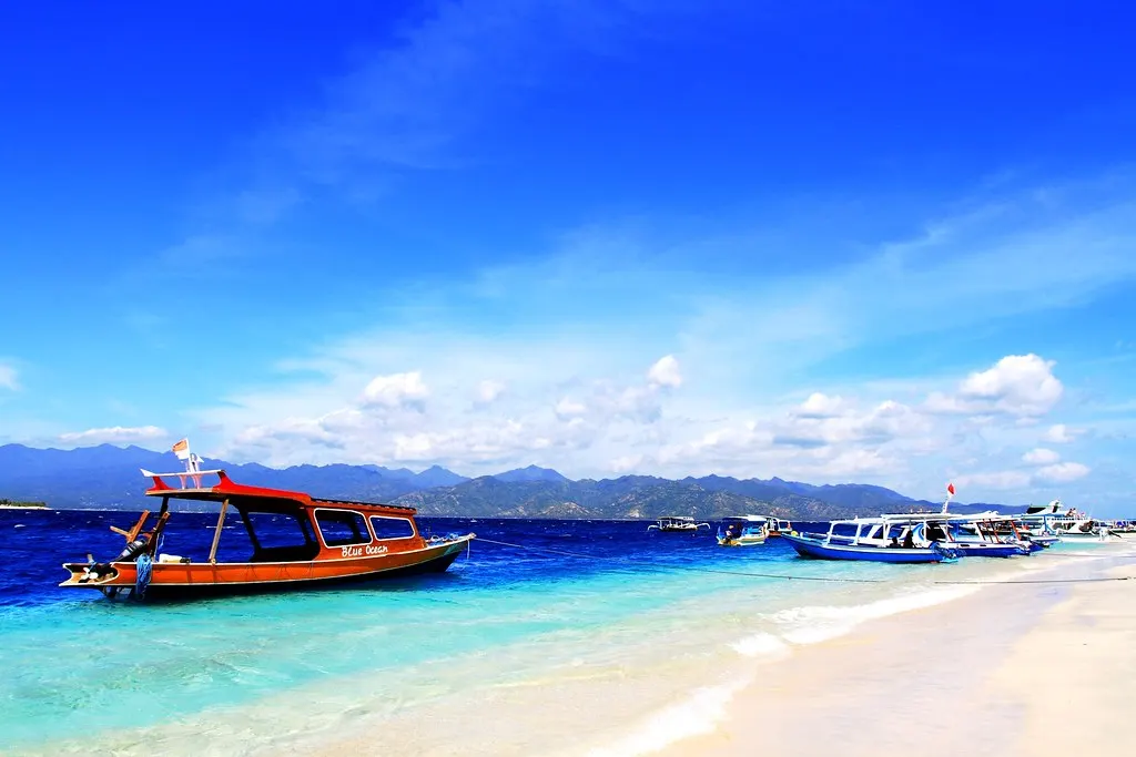 Kleurrijke boten op turquoise water bij een wit zandstrand op de Gili eilanden, met de bergen van Lombok