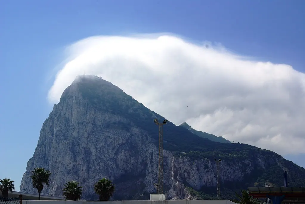 De Rots van Gibraltar met een witte wolk over de top tegen een blauwe lucht, palmbomen op de voorgrond