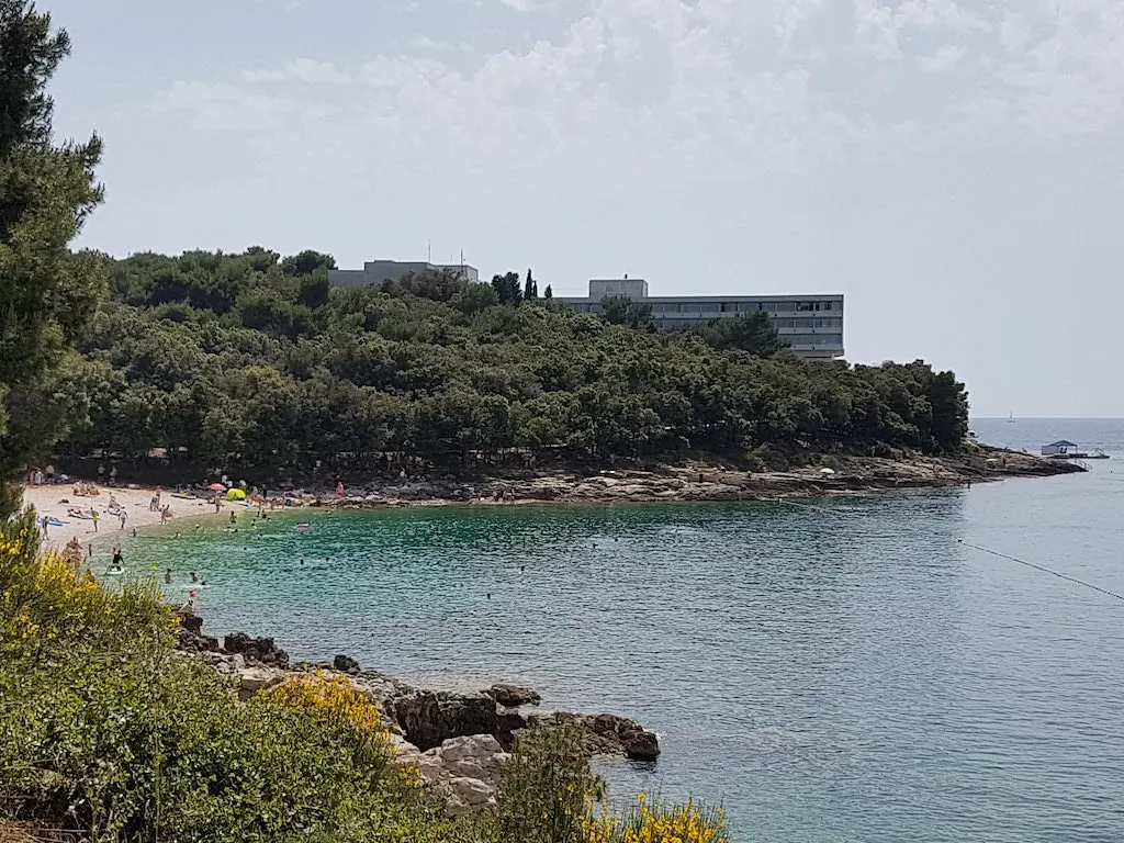 Turquoise baai met kiezelstrand omringd door dennenbos en rotsen, met een hotel op de achtergrond