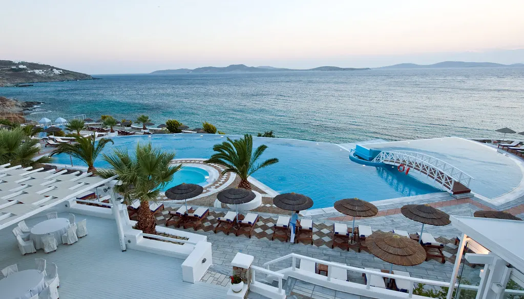 Wit hotel met infinity-zwembad, palmbomen en strooien parasols aan zee op Mykonos bij zonsondergang