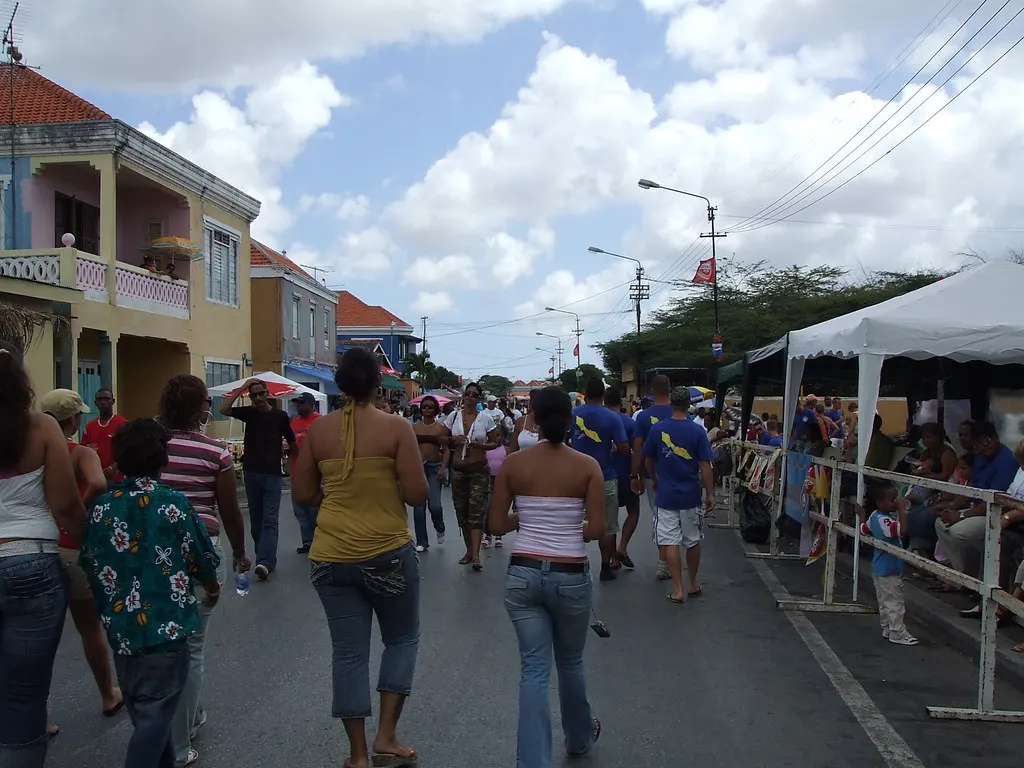 Feestgangers lopen over de route van de carnavalsoptocht tussen kleurrijke huizen en tribunes op Curaçao