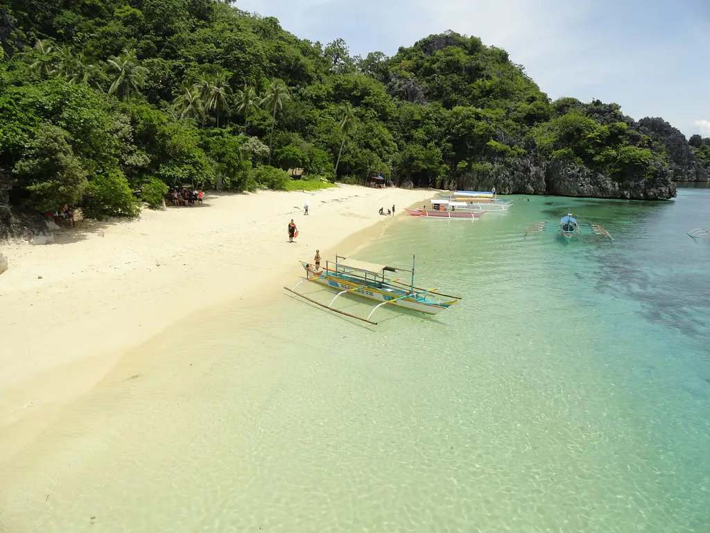 Wit zandstrand met bangka-boten aan helder turquoise water, omringd door groene kalksteenrotsen in Caramoan