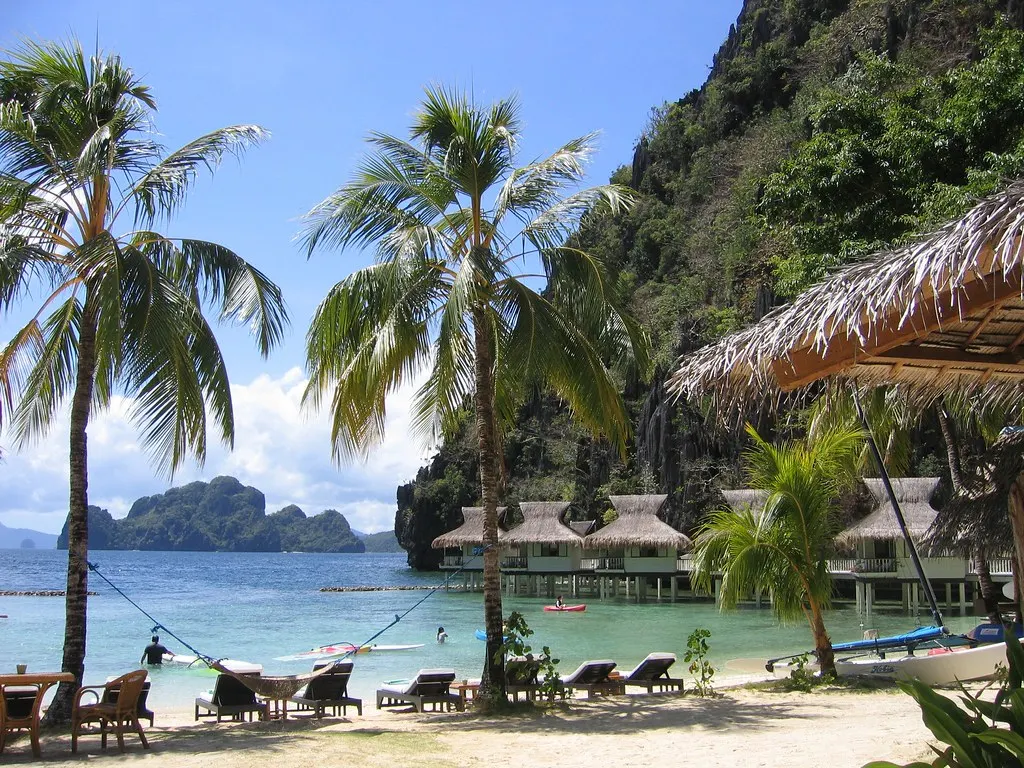Strand met palmbomen en ligstoelen, huisjes op palen boven turquoise water en kalkstenen rotsen op Miniloc Island
