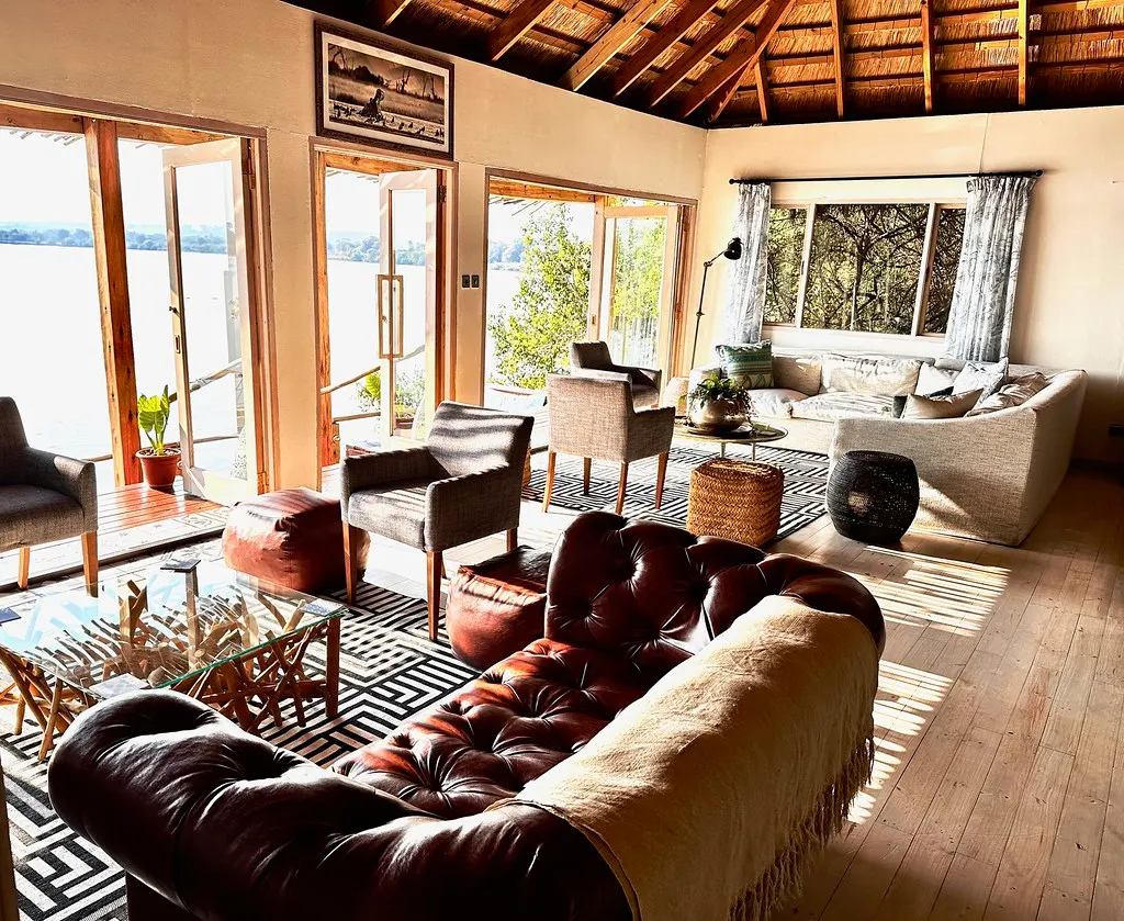 Lichte lounge met rieten dak, leren chesterfield en grote openslaande ramen met uitzicht op de Zambezi