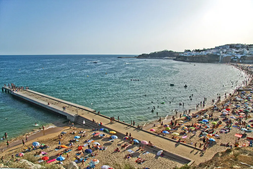 Druk strand met parasols en een lange pier in zee, met de witte huizen van Albufeira op de kliffen