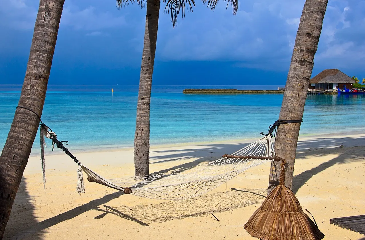 Hangmat tussen palmbomen op een wit strand met turquoise zee en een resort met waterbungalows op de Malediven