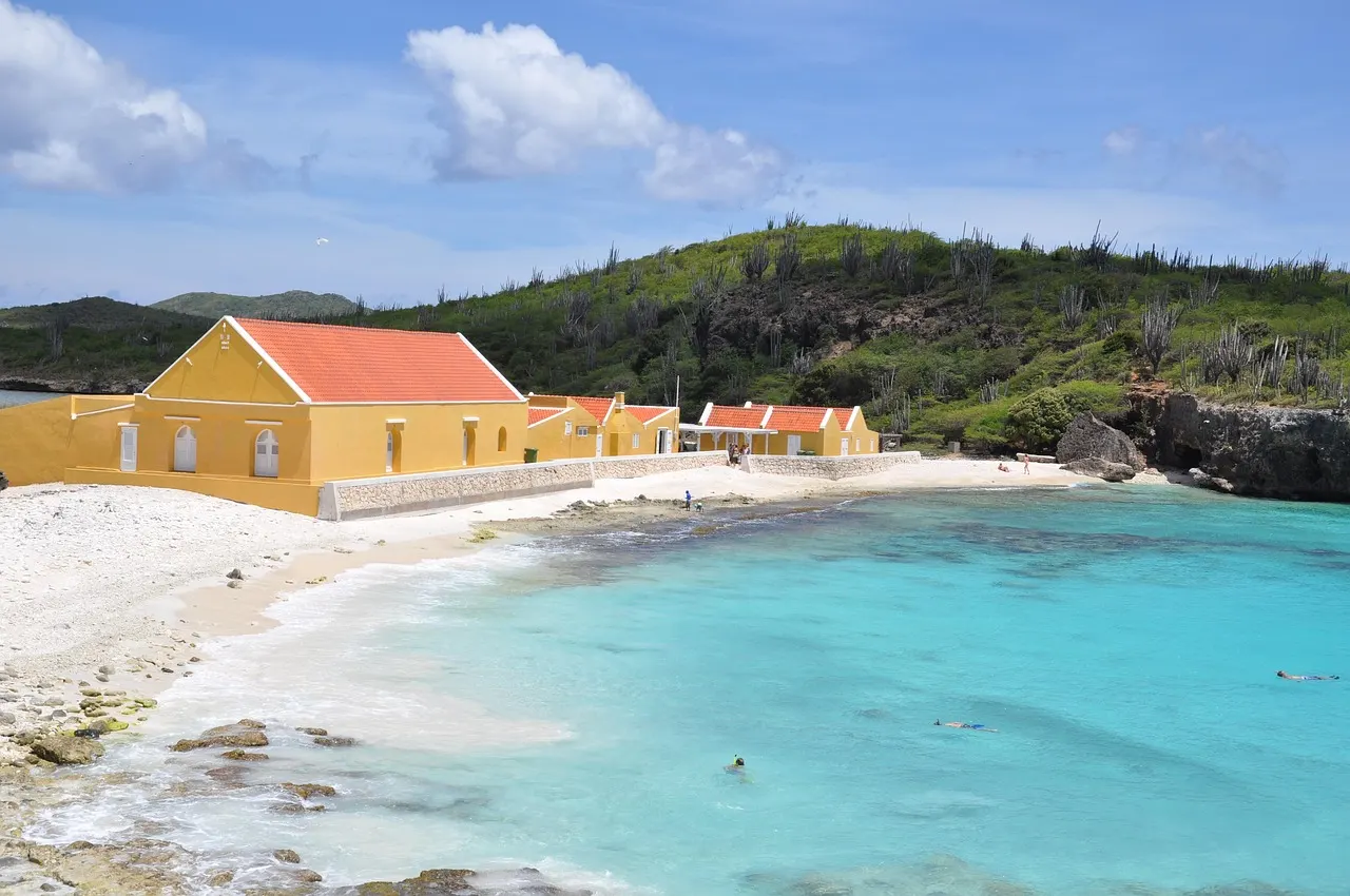 Turquoise baai met wit strand en geel gebouw met rode daken tegen een groene heuvel aan de kust van Bonaire