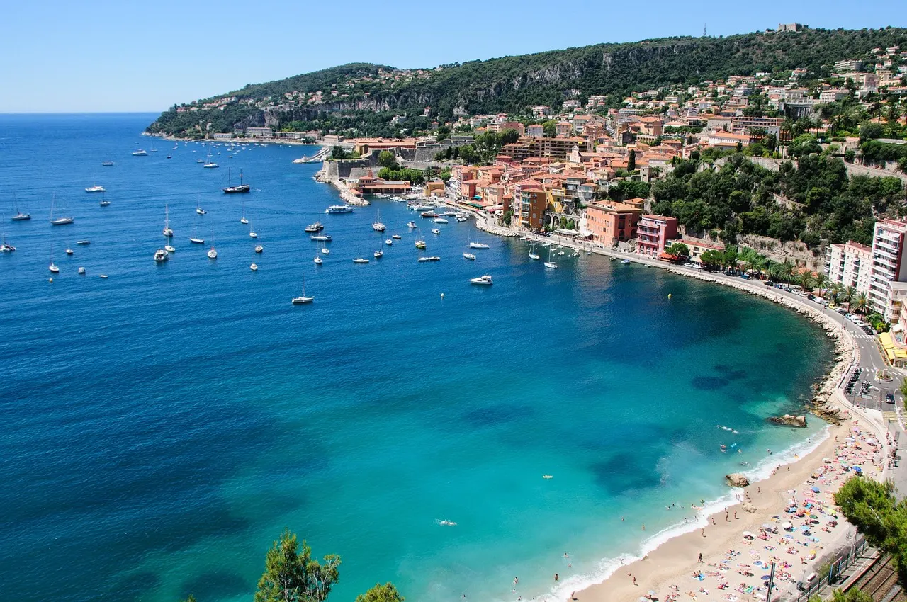 Turquoise baai van Villefranche-sur-Mer met zeilboten, strand en kleurrijke huizen tegen de groene heuvels