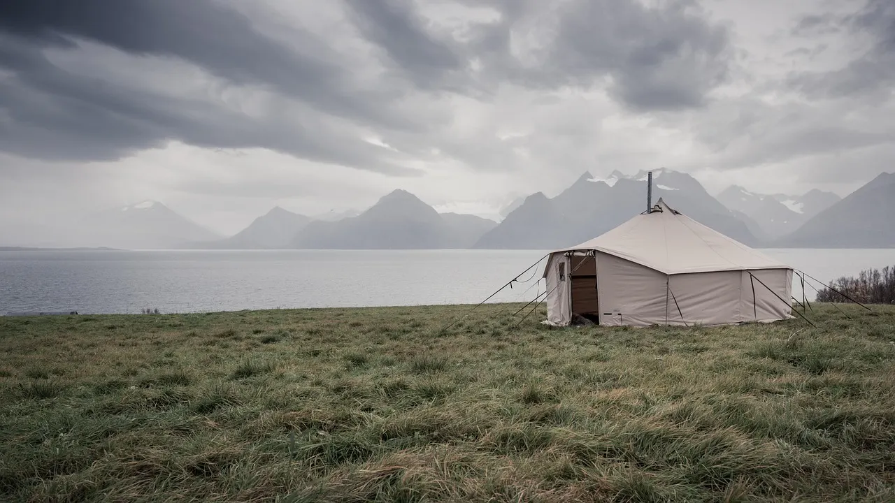 Witte canvas tent op een grasvlakte aan een meer met bergen onder een bewolkte lucht