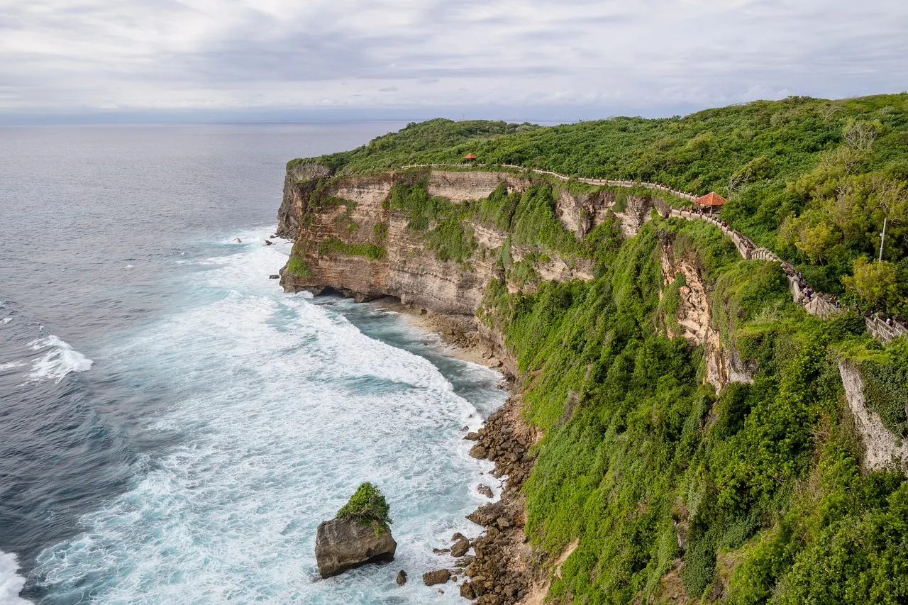 Groene kliffen met wandelpad langs de kust bij Uluwatu, met witte branding en een rots in zee