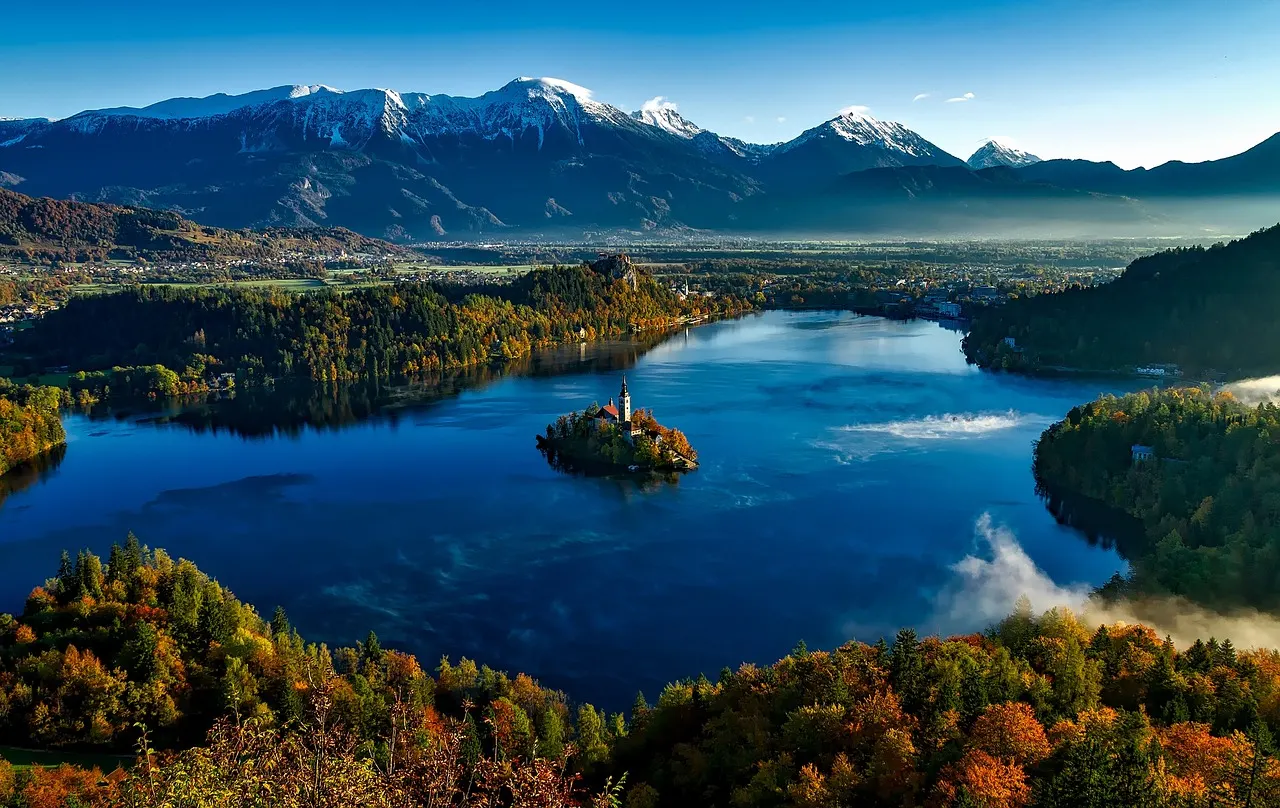Meer van Bled met het kerkeiland, herfstbossen en besneeuwde Julische Alpen onder een blauwe lucht