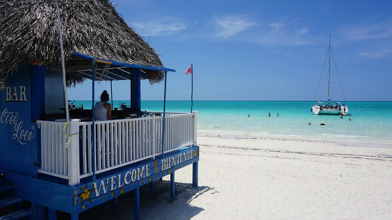Blauwe strandbar met rieten dak aan een wit strand met turquoise zee en een catamaran op Cayo Coco
