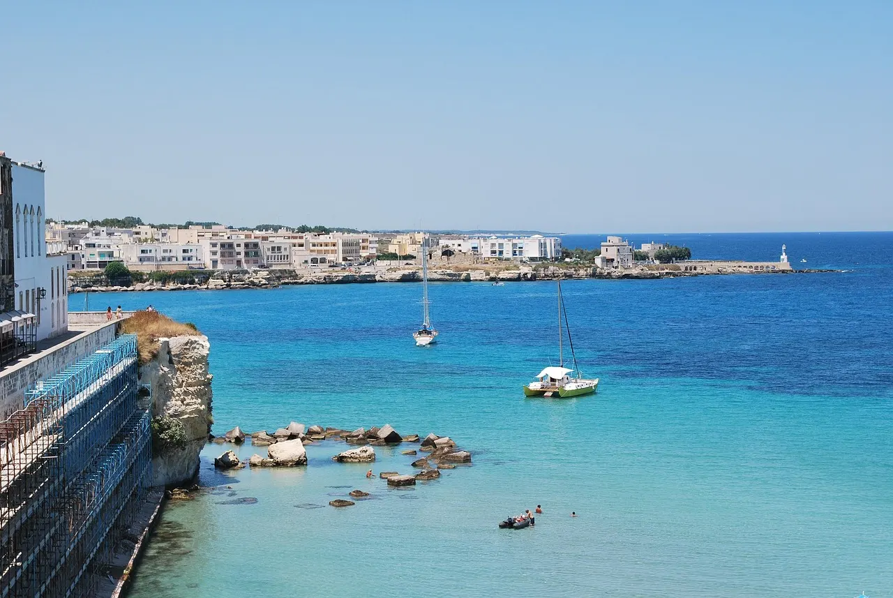 Turquoise baai bij Otranto met rotsblokken in het heldere water, zeilboten en witte huizen langs de kust