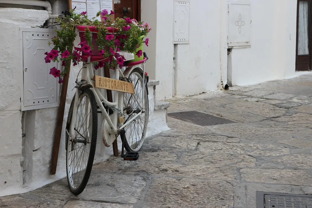 Witte fiets met roze bloemen als restaurantbord in een wit gekalkt straatje met kasseien in Ostuni