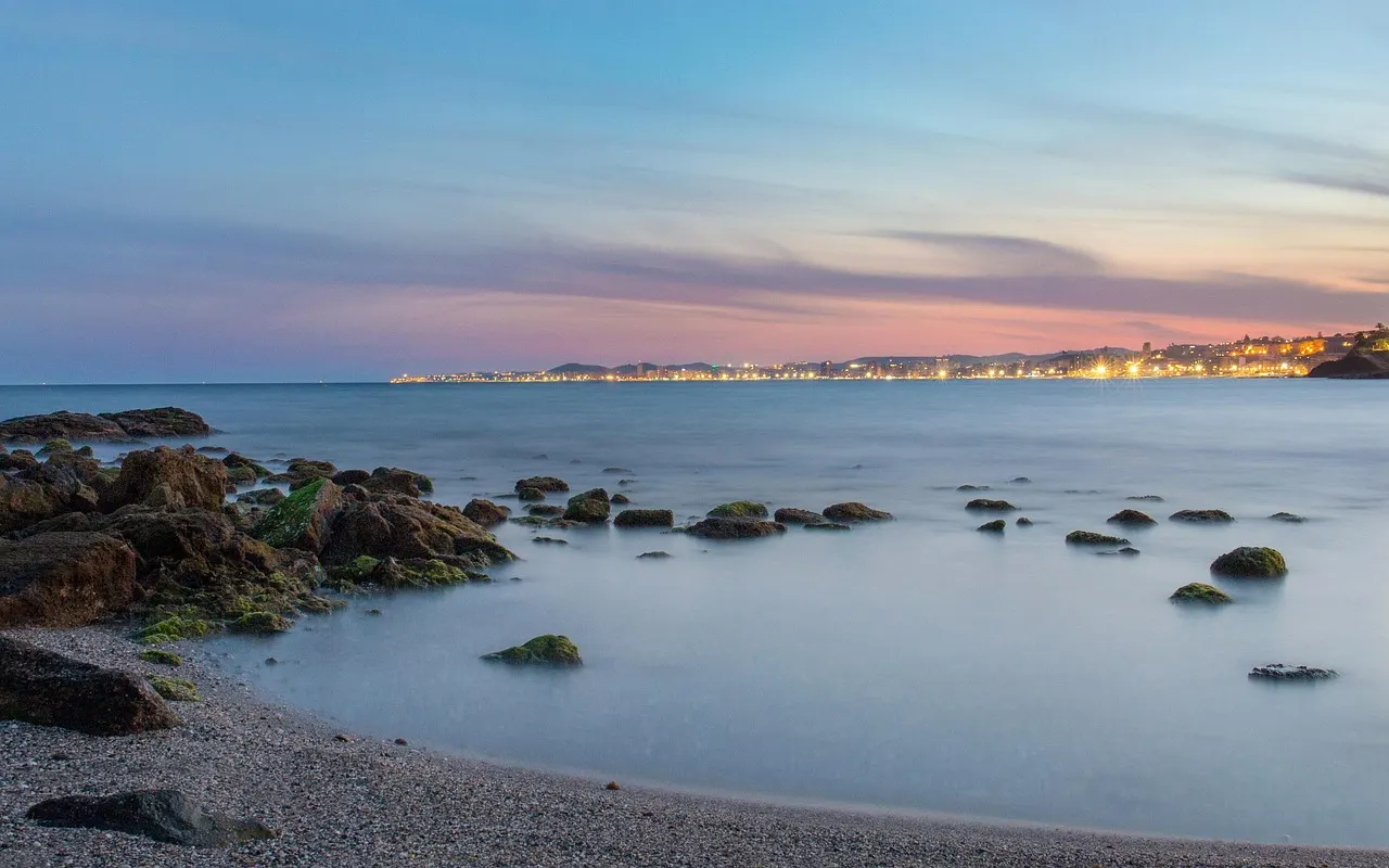 Kiezelstrand met rotsen in kalme zee bij schemering, met verlichte kustplaats aan de horizon