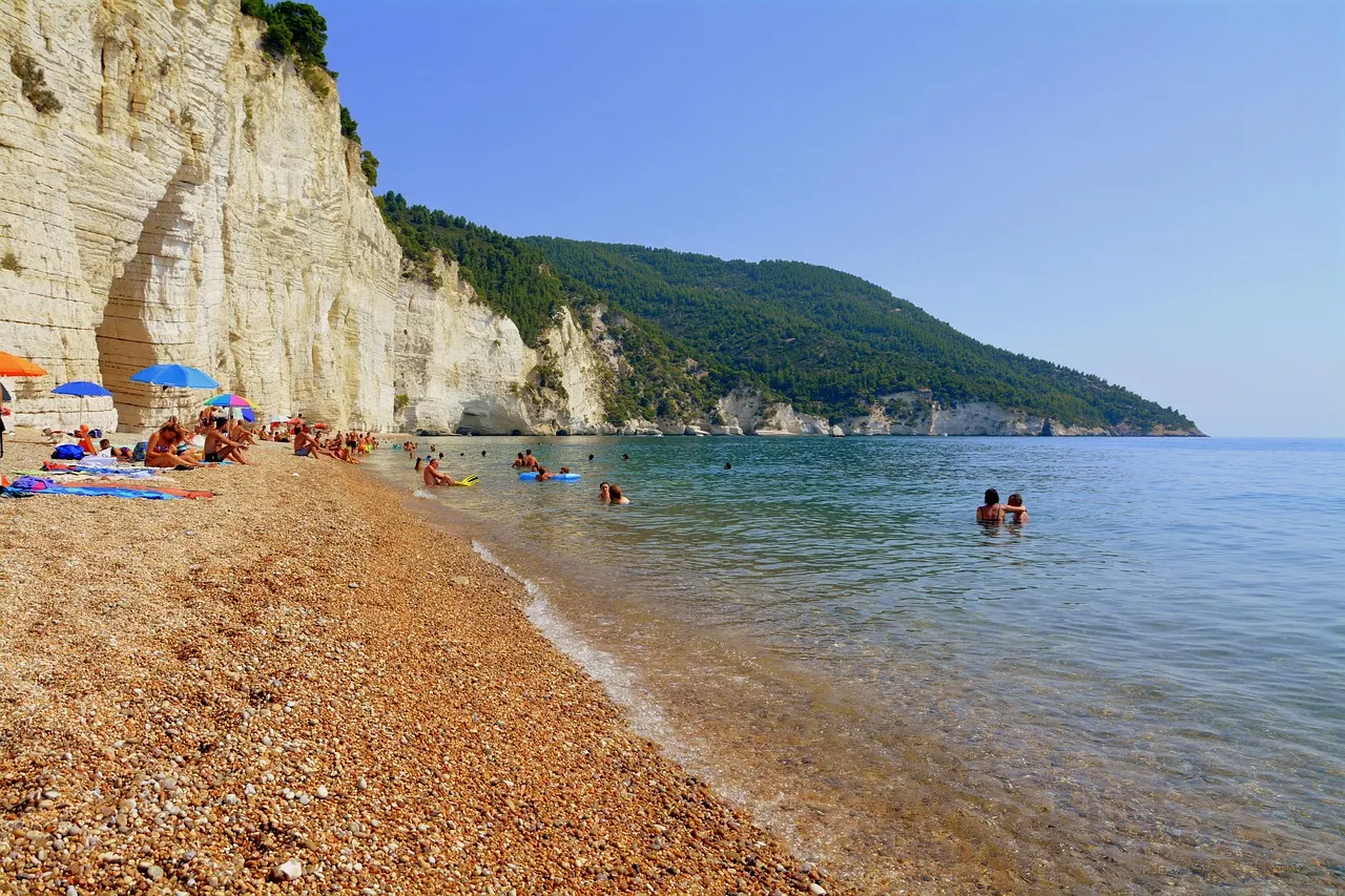 Kiezelstrand onder hoge witte kalkrotsen en beboste heuvels aan helderblauwe zee in Gargano