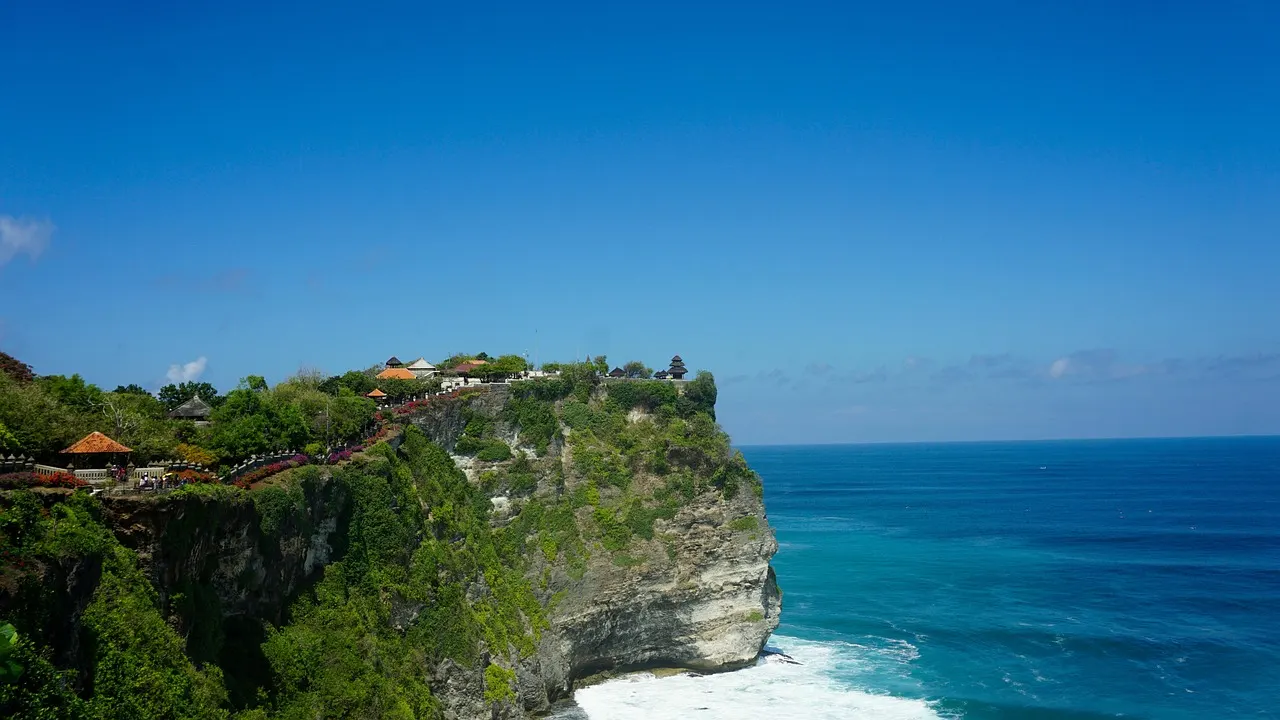 Kliffen van Uluwatu met tempel bovenop, turquoise oceaan en branding onder een strakblauwe lucht