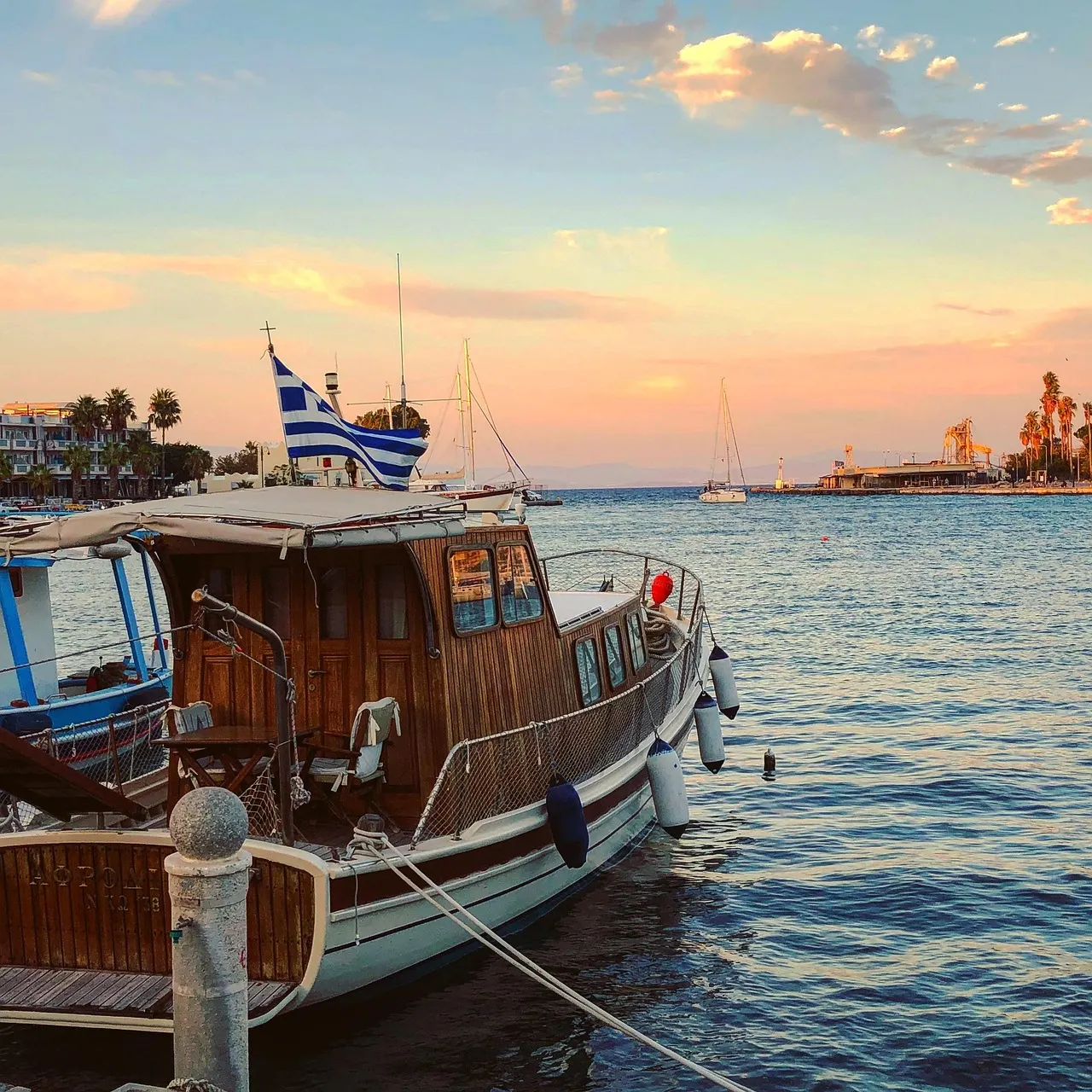 Houten boot met Griekse vlag in de haven van Kos bij zonsondergang, met palmbomen aan de kade