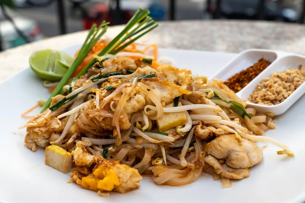 Bord Pad Thai met kip, tofu, taugé, bieslook en limoen, met gemalen pinda's en chilivlokken ernaast