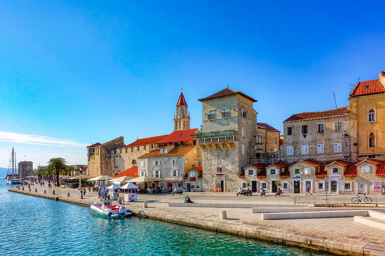 Zonnige kade van Trogir met stenen huizen, kerktoren, palmbomen en turquoise water met bootjes