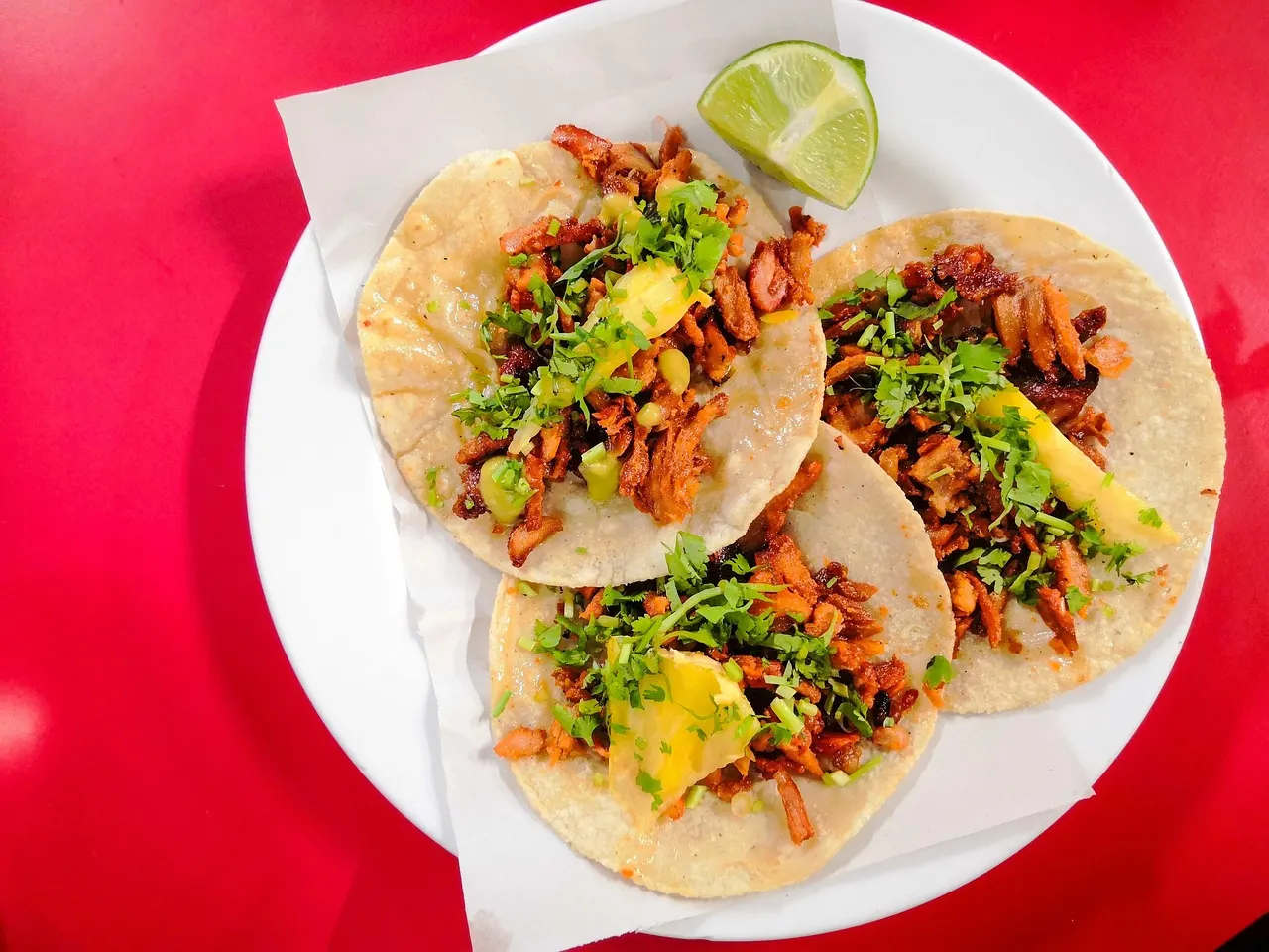 Bord met drie tacos al pastor met koriander, ananas en een partje limoen op een rode tafel