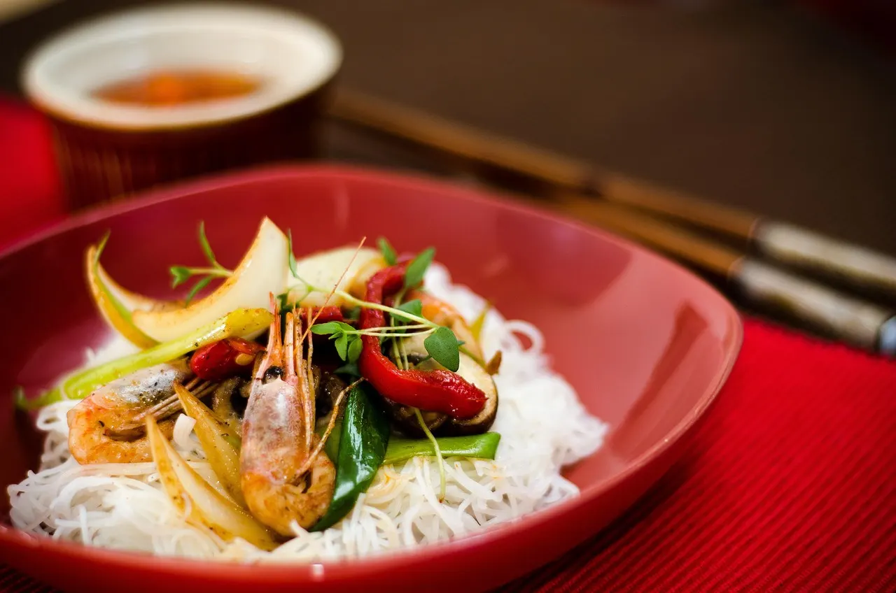 Rijstnoedels met garnalen, paprika en groenten in een rode schaal met stokjes op de achtergrond