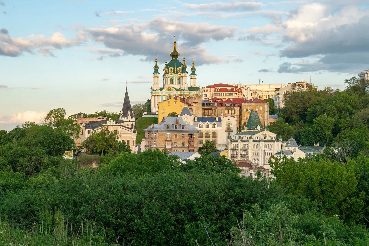Sint-Andreaskerk boven de historische huizen van Kiev, omringd door groen onder een zachte avondlucht