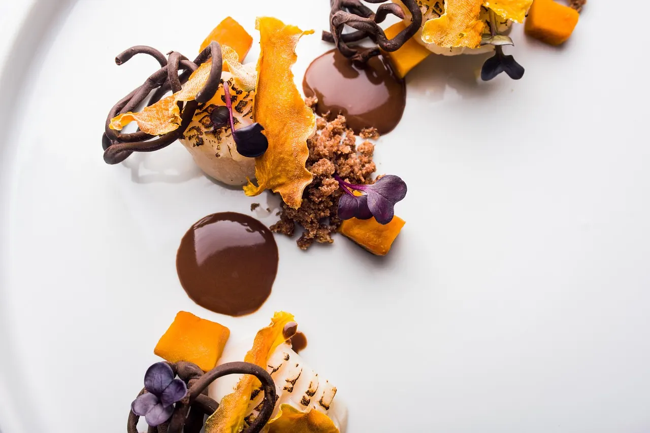Verfijnd fine-dining dessert met chocolade, oranje blokjes, krokante garnering en paarse bloemetjes op een wit bord
