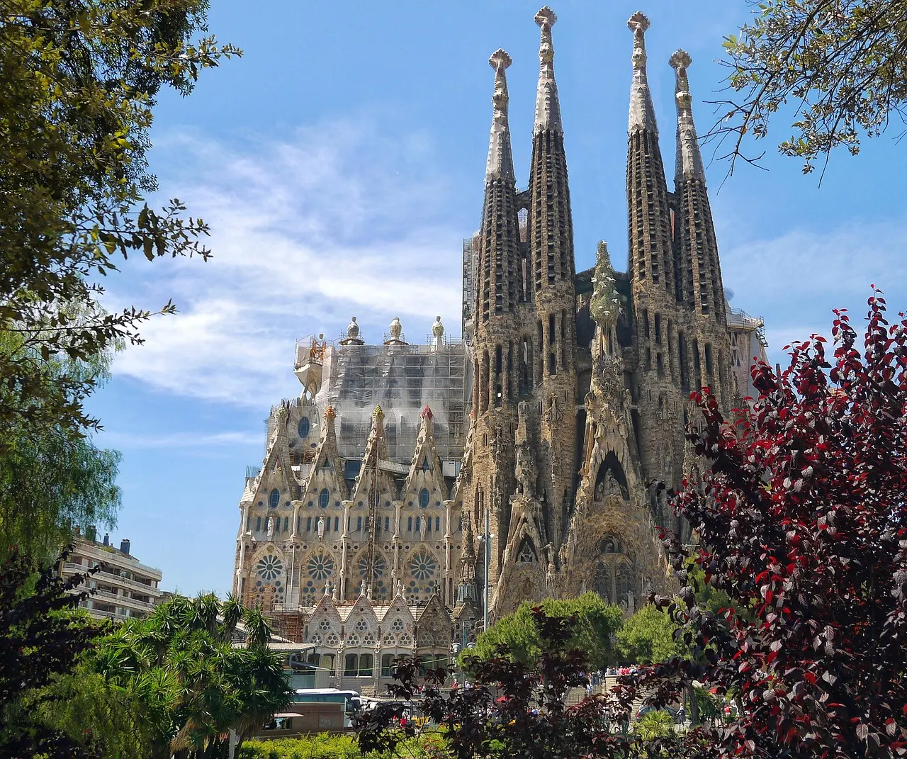 De torens van de Sagrada Família in Barcelona onder een blauwe lucht, omlijst door groene en rode bomen