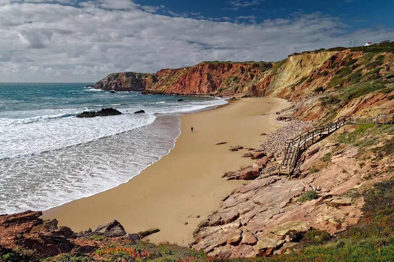 Goudgeel strand tussen rode kliffen met houten trap en golven aan de kust van de Algarve