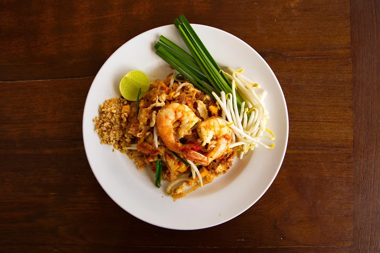 Bord Pad Thai met garnalen, taugé, bieslook, gemalen pinda's en een partje limoen op een houten tafel