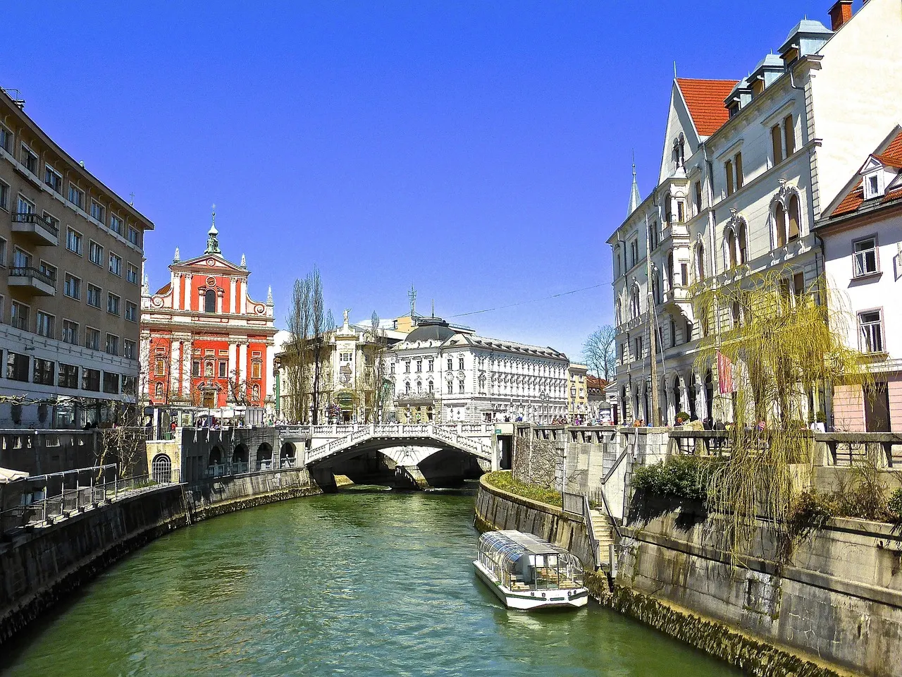 De rivier de Ljubljanica met de Drievoudige Brug, een rondvaartboot en de roze Franciscanerkerk onder blauwe lucht