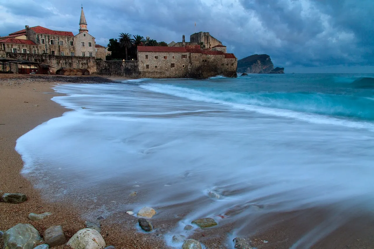 De ommuurde oude stad van Budva aan het strand, met golven en het eiland Sveti Nikola onder een bewolkte lucht