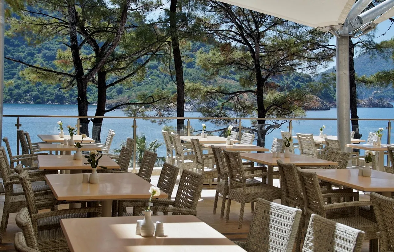 Terras van een restaurant met rieten stoelen en houten tafels onder pijnbomen, met uitzicht op zee