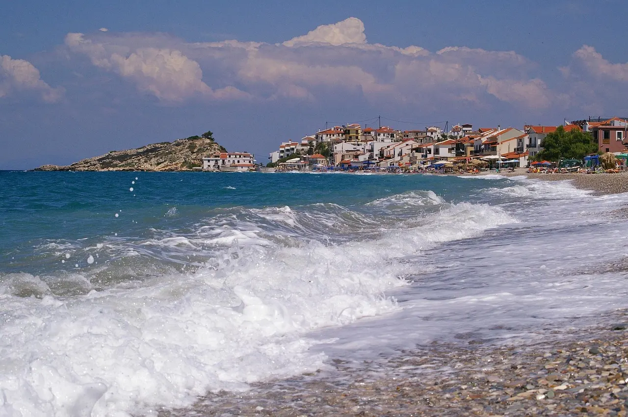Golven op een kiezelstrand met witte huizen en een rotsachtig eilandje op de achtergrond, Samos