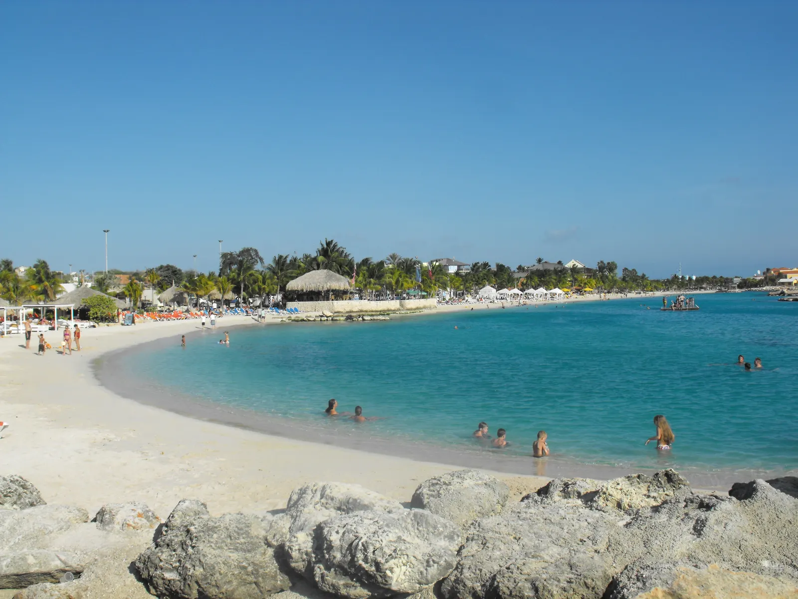 Wit zandstrand met turquoise baai, palmbomen en een rieten parasol, met rotsen op de voorgrond