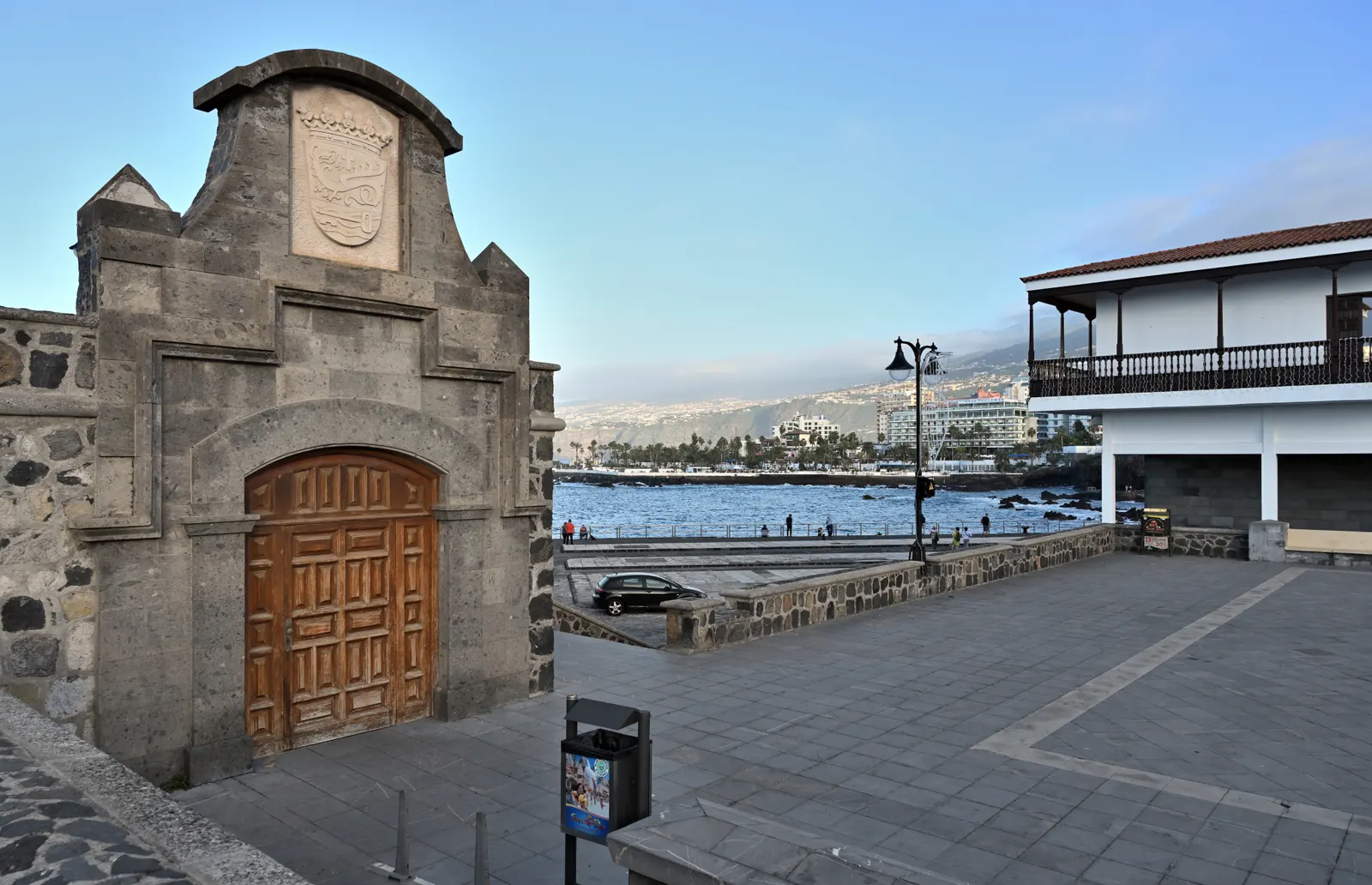 Historische stenen poort met houten deur aan de kade van Puerto de la Cruz, met zee en kustlijn op de achtergrond