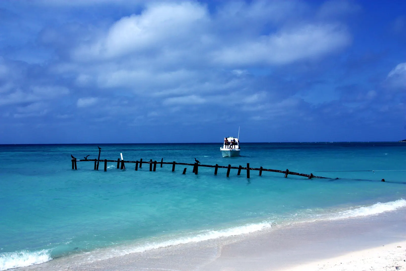 Wit zandstrand met turquoise zee, een oude houten steiger en een bootje onder een blauwe lucht met wolken