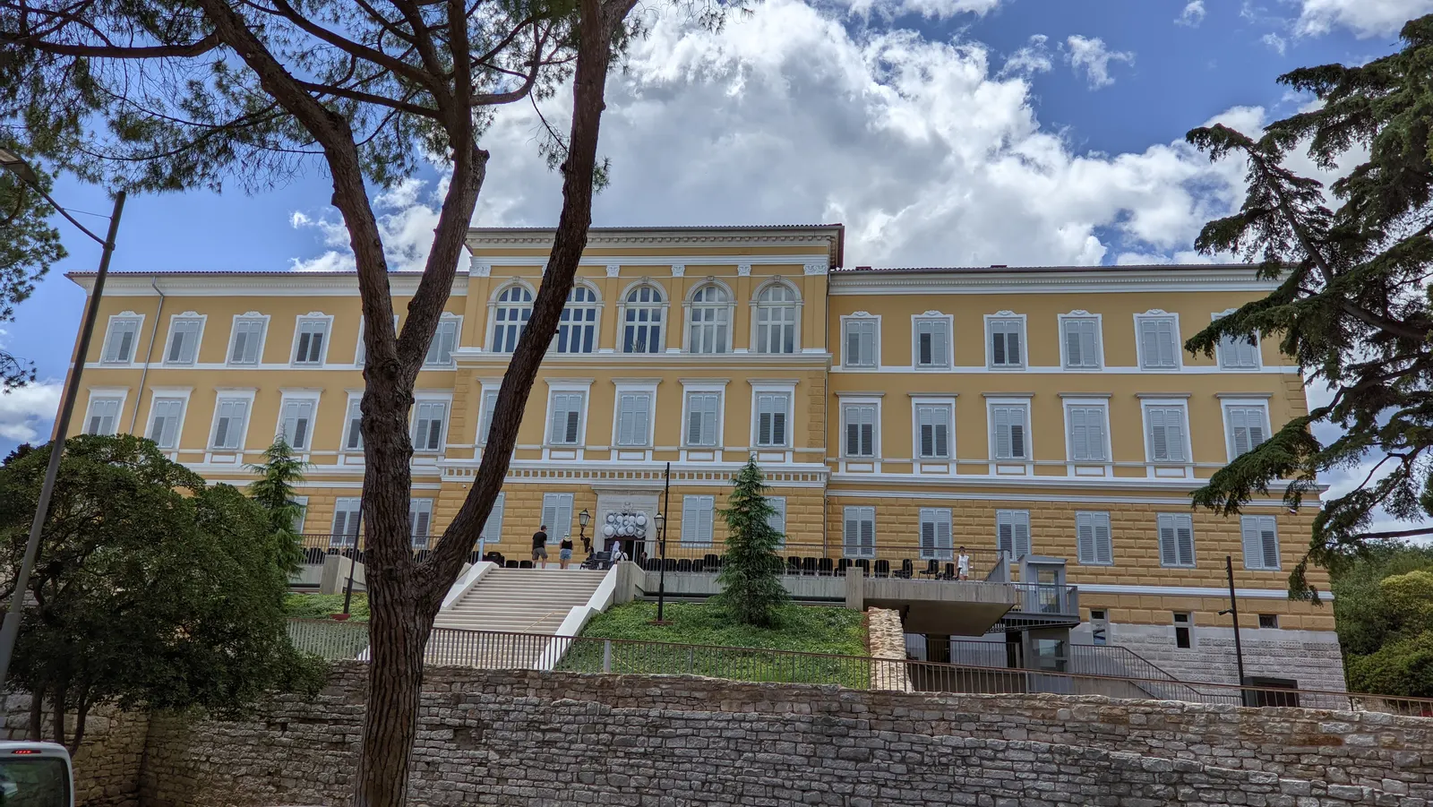 Geel museumgebouw van het Archeologisch Museum van Istrië in Pula met trappen, bomen en blauwe lucht