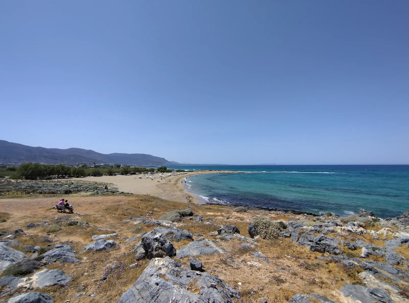Lang zandstrand met turquoise zee, rotsachtige kust op de voorgrond en bergen op de achtergrond